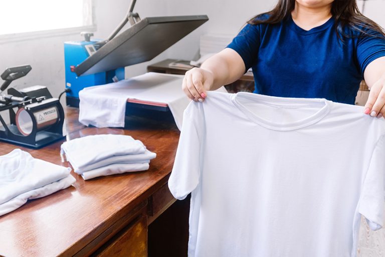 Female holding a white T-shirt next to the cloths printing machine and T-shirts on the wooden table